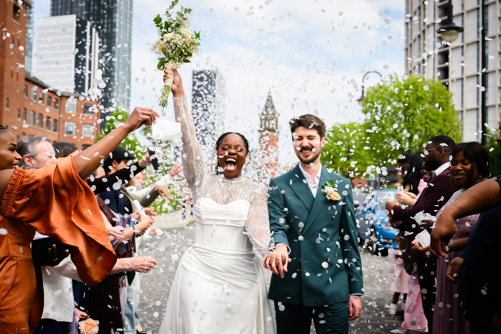 The bride and groom walking through a confetti shower outside The Castlefield Rooms in Manchester, with the city skyline visible in the background, the bride raising her bouquet in celebration.