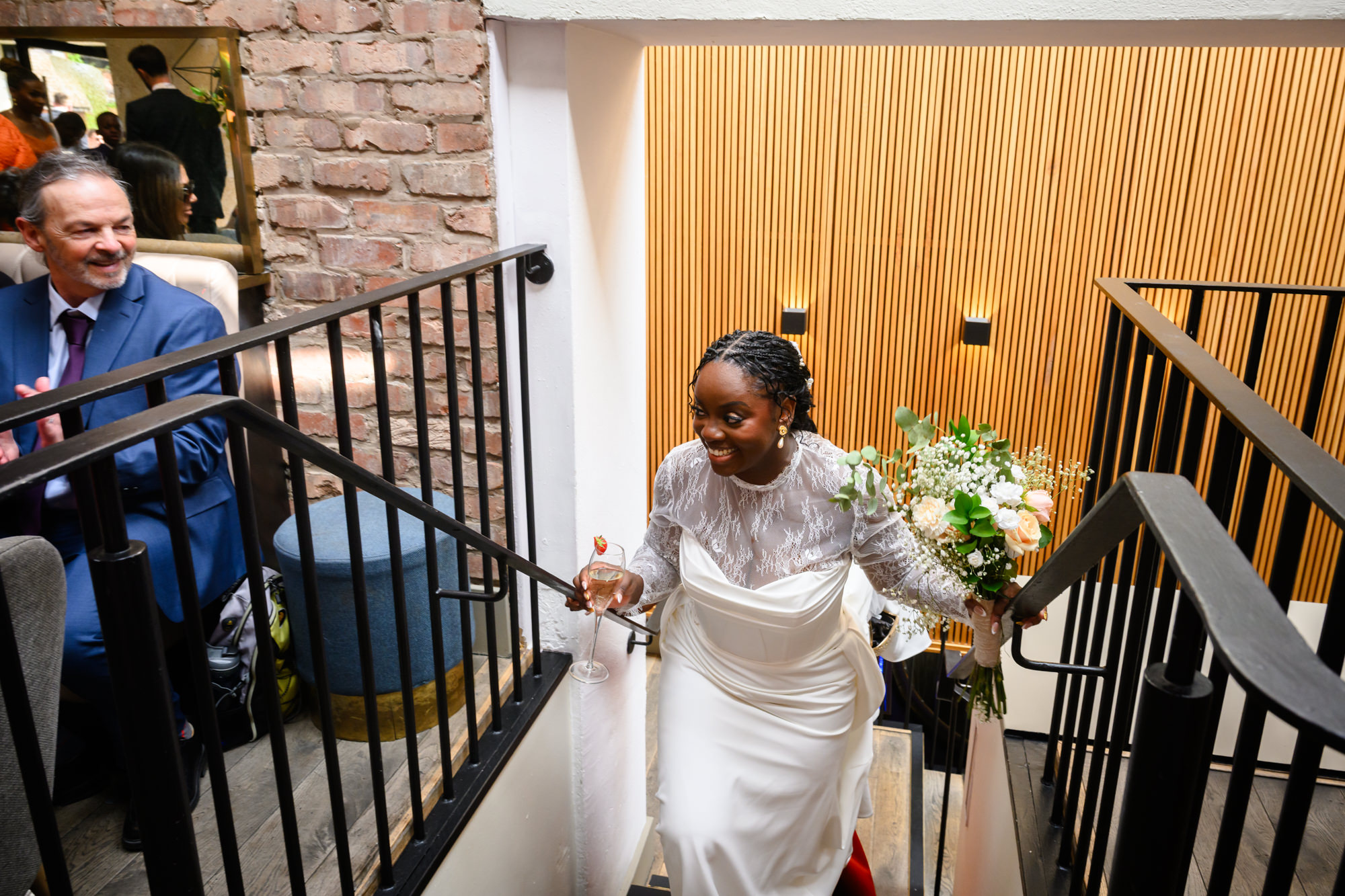 The bride walking up the stairs at The Castlefield Rooms during the drinks reception, holding her bouquet and a glass of prosecco, smiling.