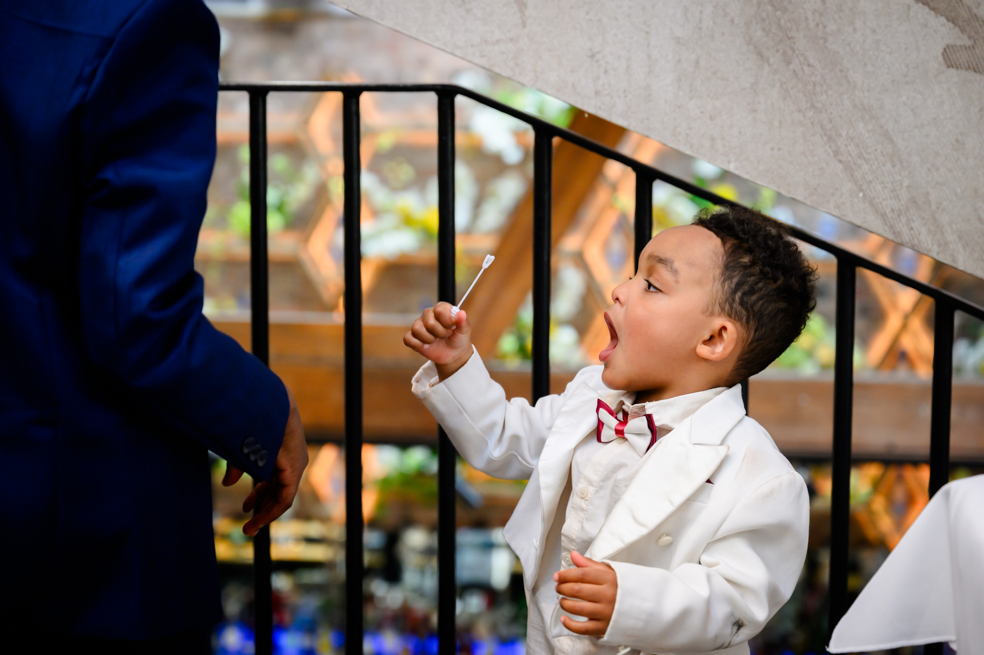 A young wedding guest in a white suit and red bow tie blowing bubbles on the stairs at The Castlefield Rooms during the drinks reception.