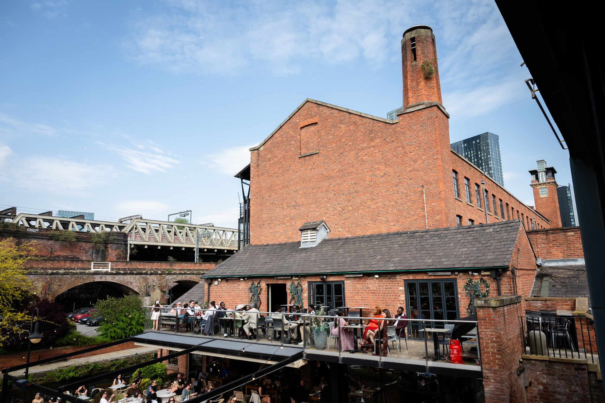 Wide shot of wedding guests enjoying the drinks reception on the balcony at The Castlefield Rooms, with the Castlefield viaduct, canal and Manchester city skyline visible in the background.