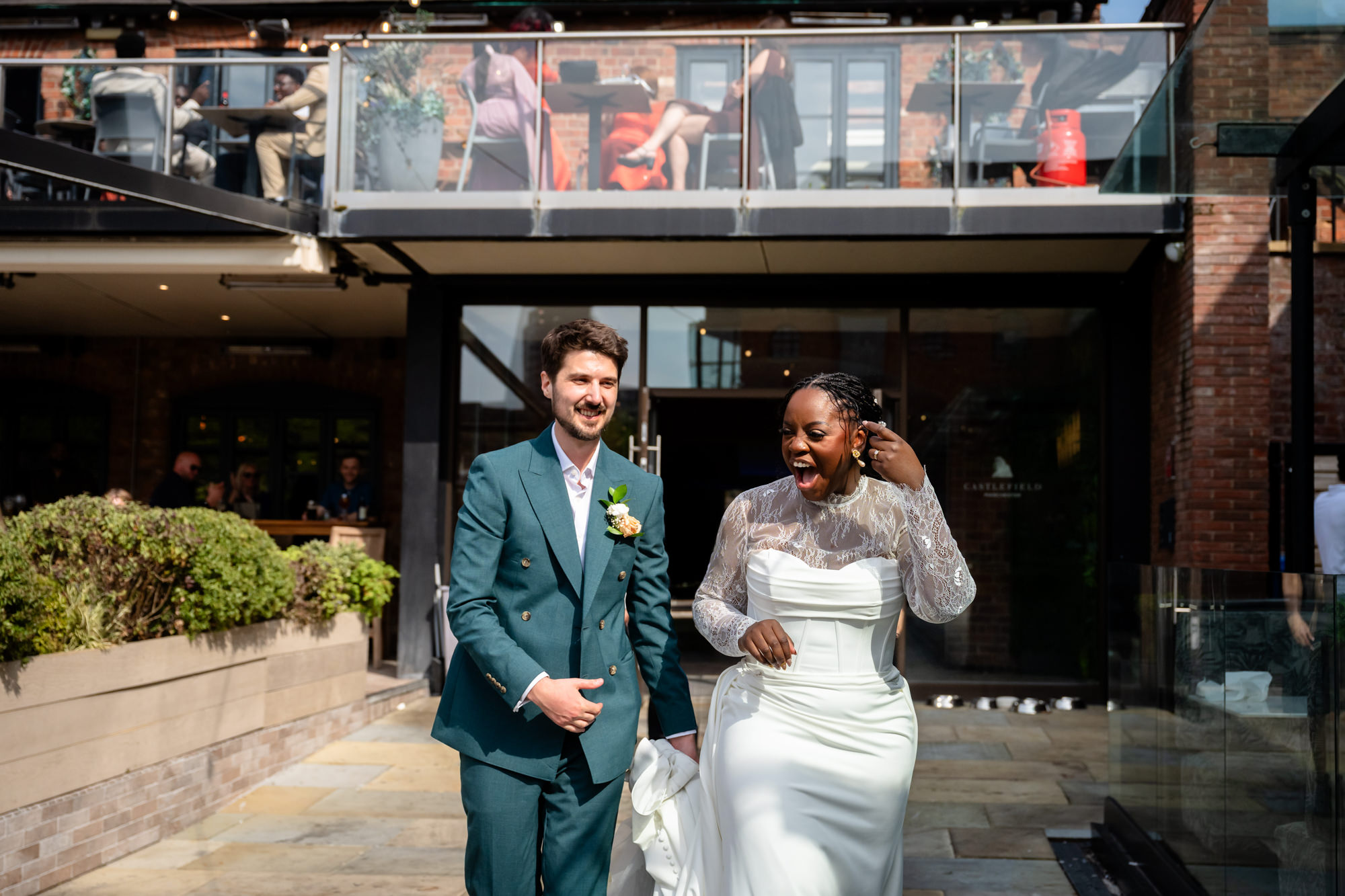 The bride and groom laughing together outside The Castlefield Rooms during their wedding portrait session, with guests visible on the balcony above.