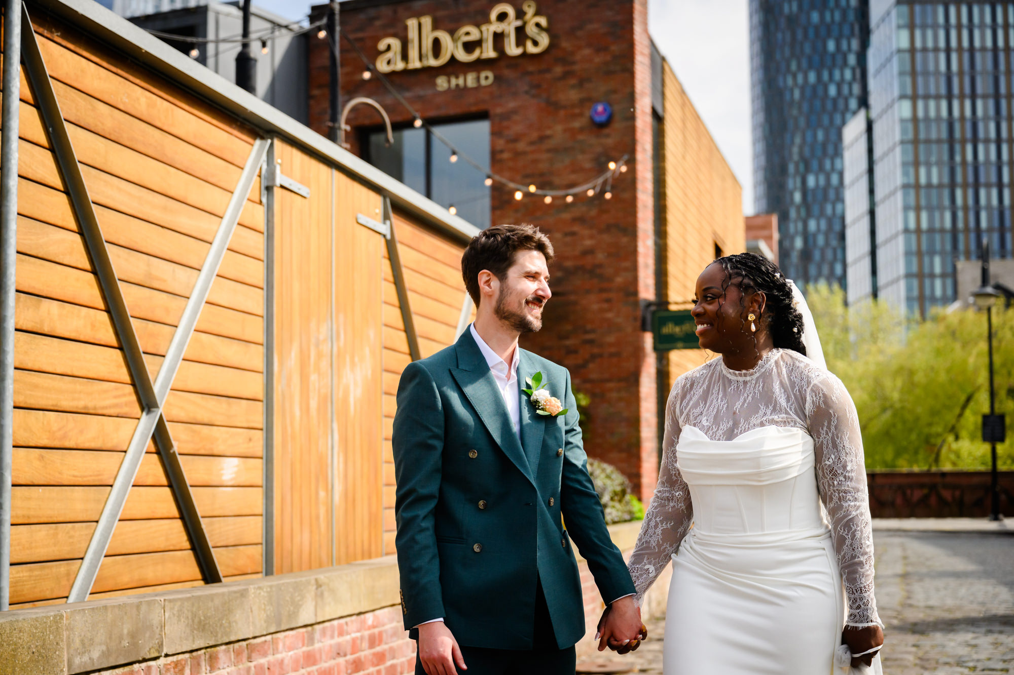 The bride and groom walking hand in hand past Albert's Shed in Castlefield during their wedding portrait session, with the Manchester city skyline in the background.