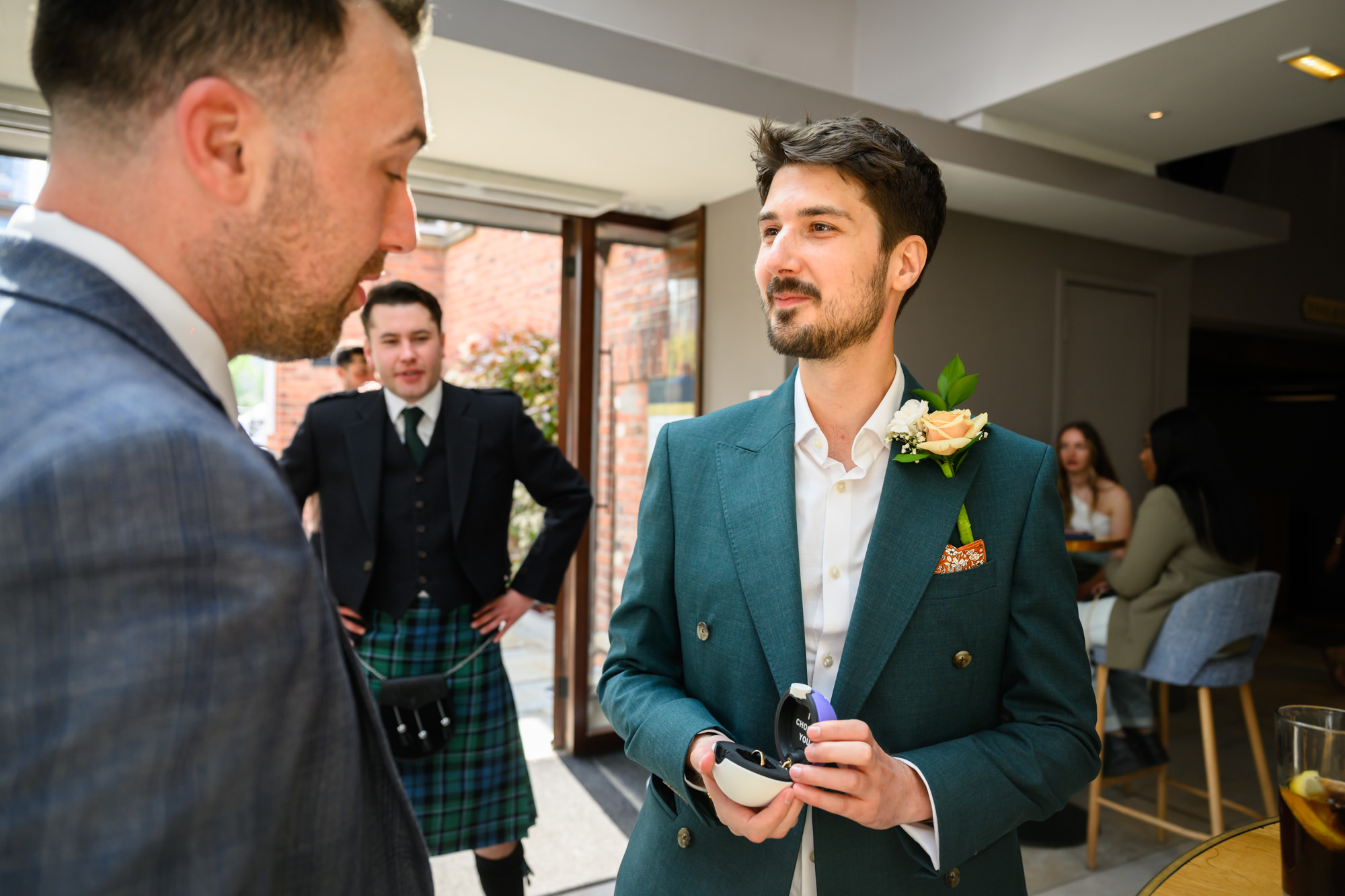 The groom in a teal suit holding the custom pokeball ring boxes while chatting with a guest at The Castlefield Rooms, with a guest in a kilt visible in the background.