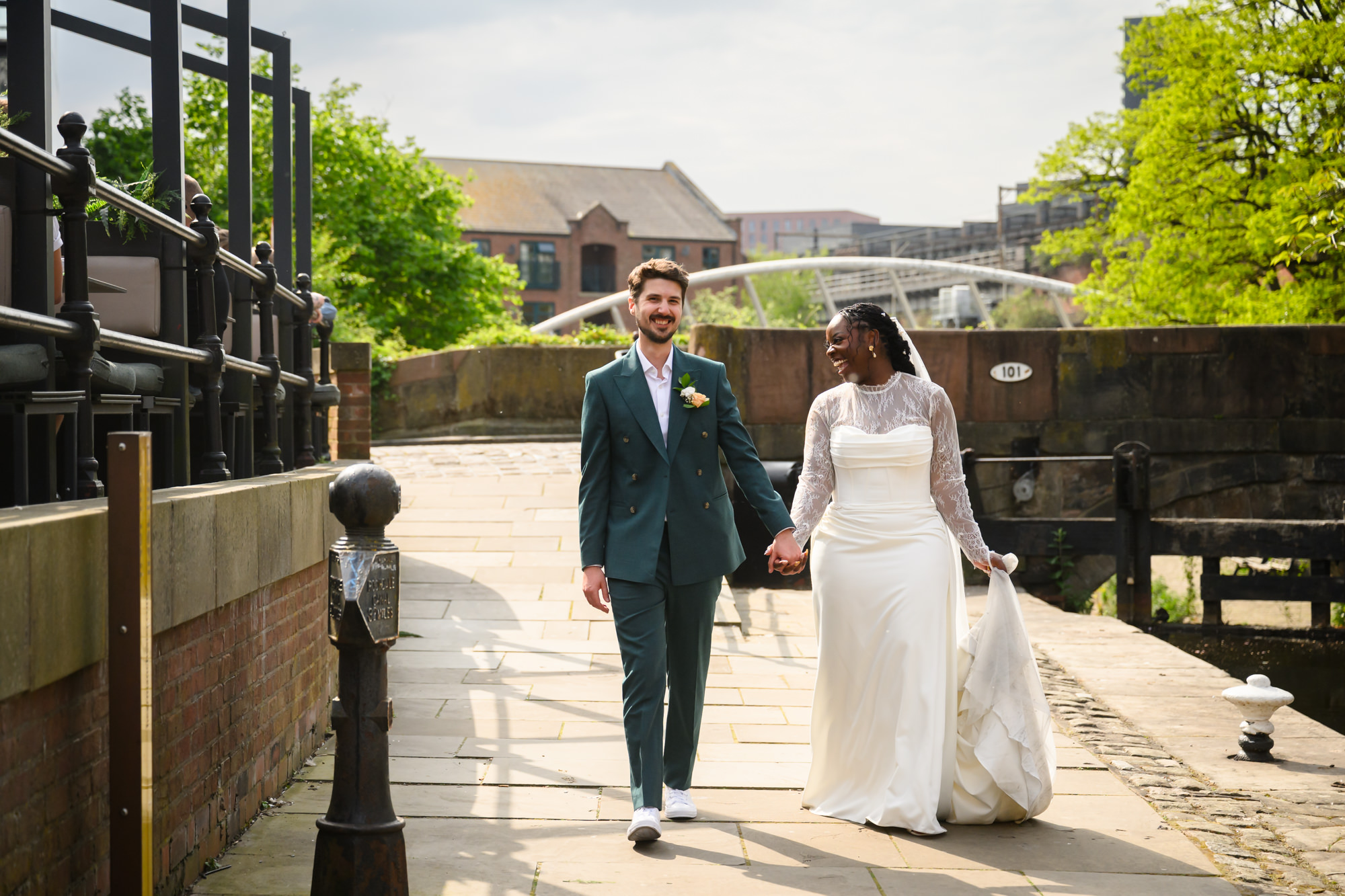 The bride and groom walking hand in hand along the Bridgewater Canal during their wedding portrait session, with a footbridge and canal boats visible in the background.