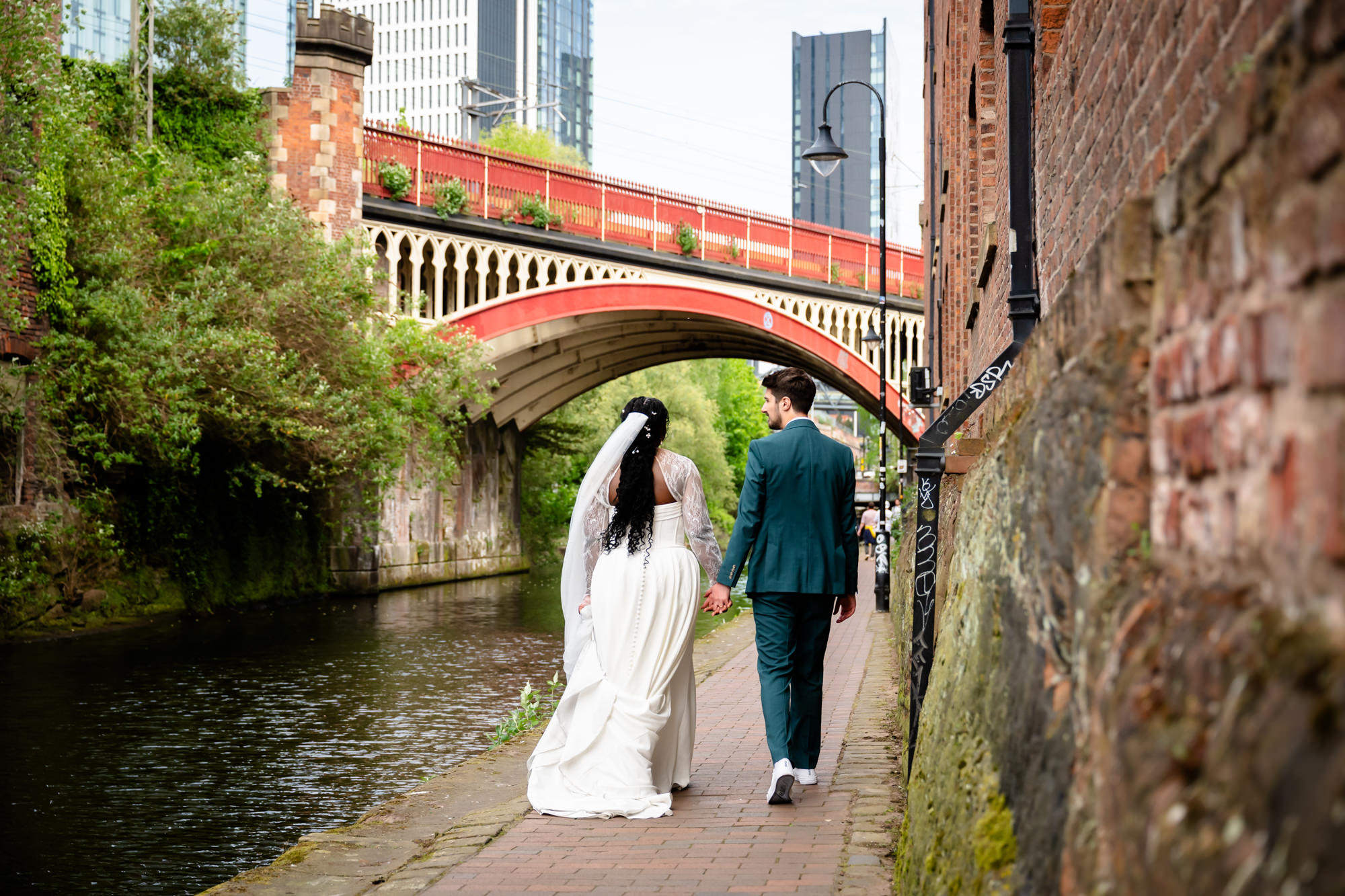 The bride and groom walking hand in hand along the Bridgewater Canal towpath during their wedding portrait session, with the iconic red Castlefield railway viaduct visible behind them.
