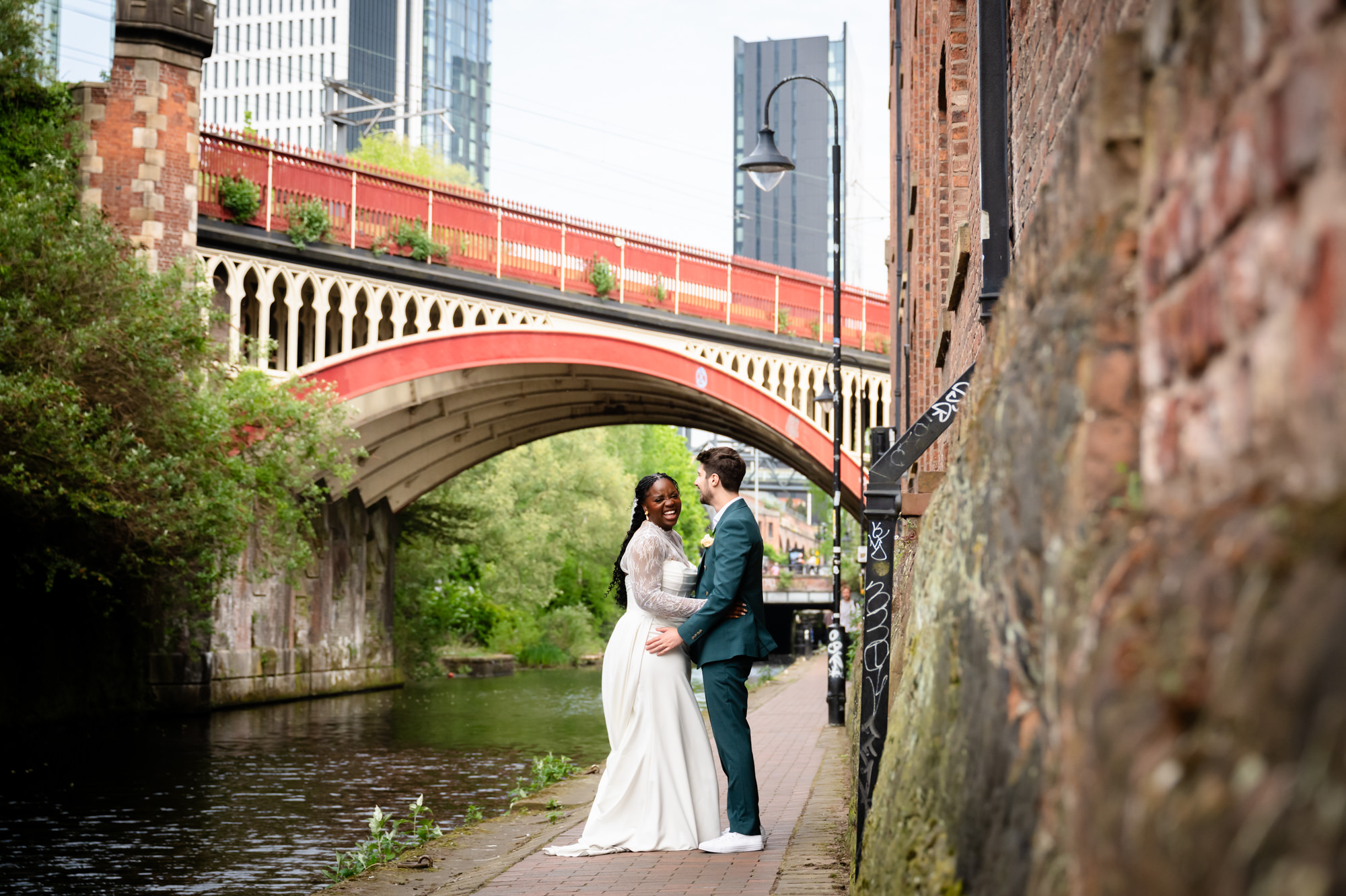 The bride and groom laughing together on the Bridgewater Canal towpath during their wedding portrait session, with the red Castlefield railway viaduct in the background.