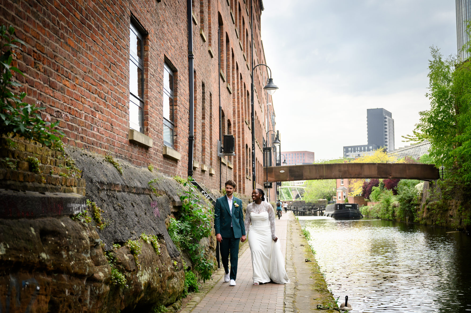 The bride and groom walking along the Bridgewater Canal towpath during their wedding portrait session, with a canal narrowboat and the Manchester skyline visible in the background.