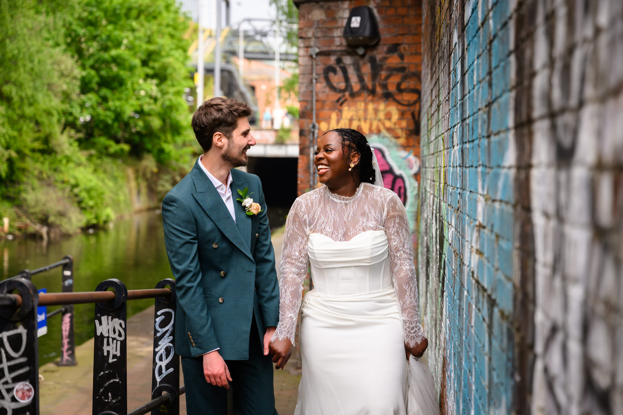 The bride and groom laughing together against a graffiti wall on the Bridgewater Canal towpath during their wedding portrait session in Castlefield, Manchester.