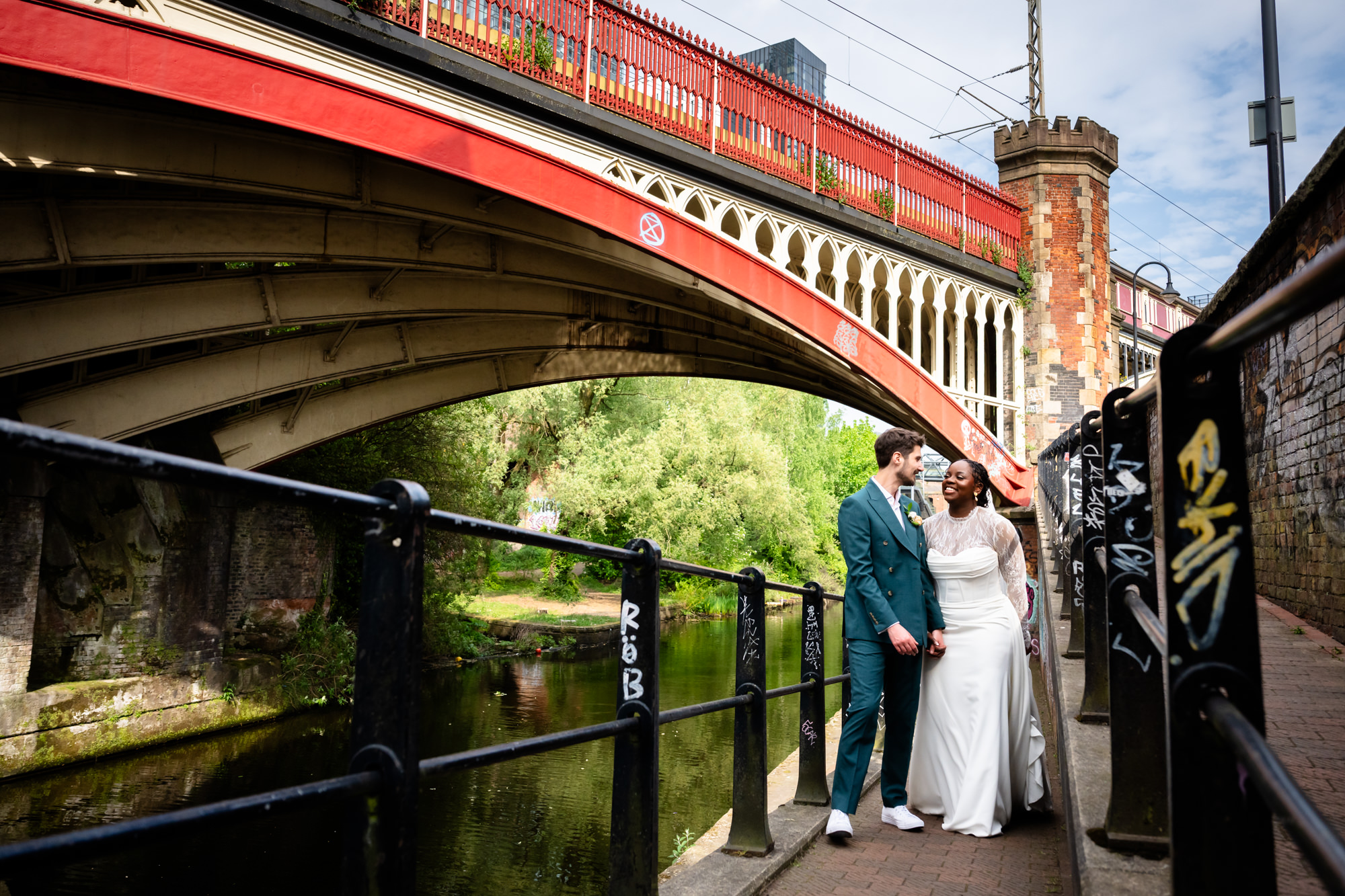 The bride and groom standing together under the red Castlefield railway viaduct during their wedding portrait session along the Bridgewater Canal in Manchester.