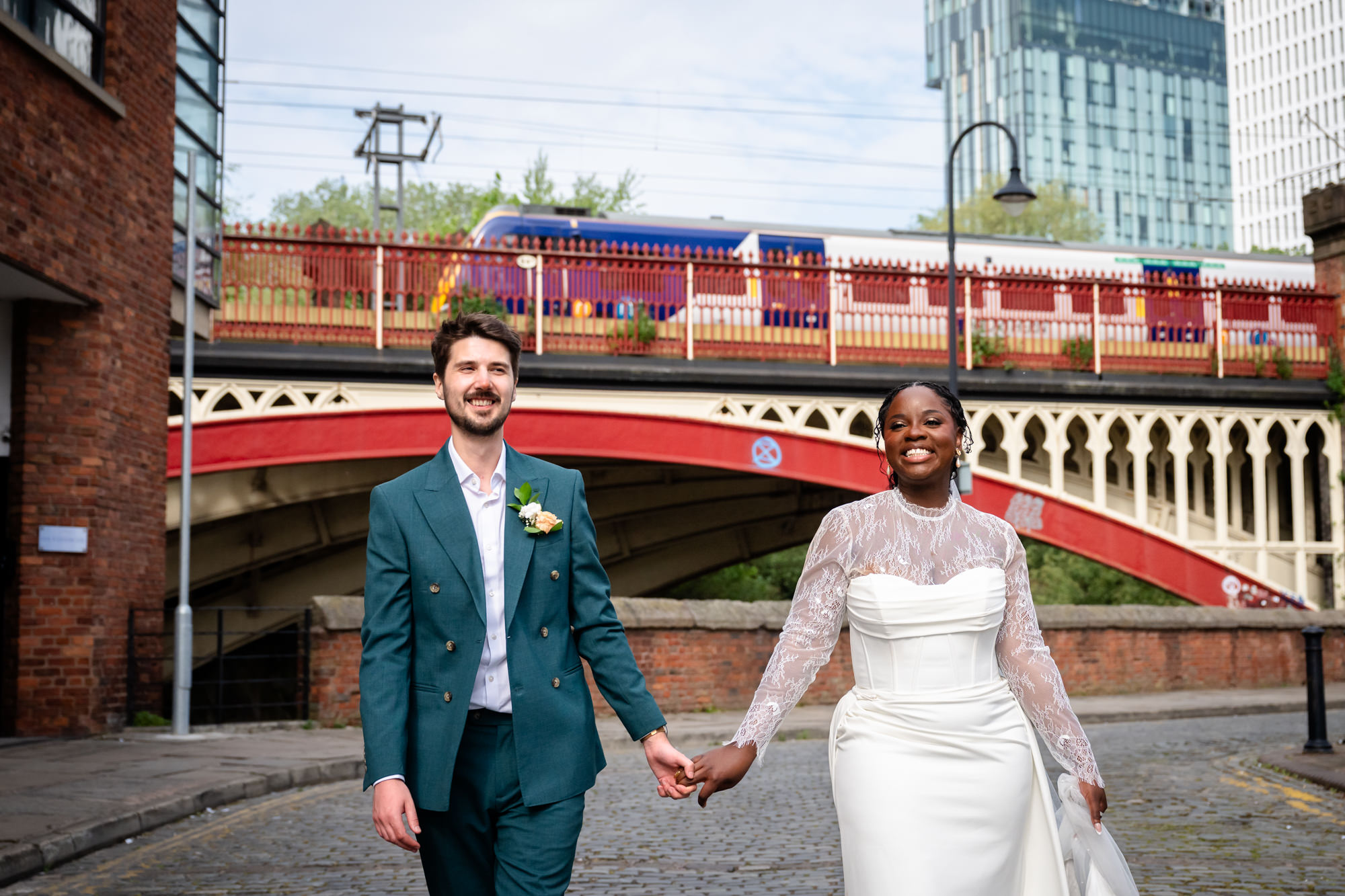 The bride and groom walking hand in hand on the cobbled streets of Castlefield during their wedding portrait session, with the red railway viaduct and a passing train visible in the background.
