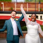 The bride and groom celebrating with arms raised on the cobbled streets of Castlefield during their wedding portrait session, with the red railway viaduct behind them.