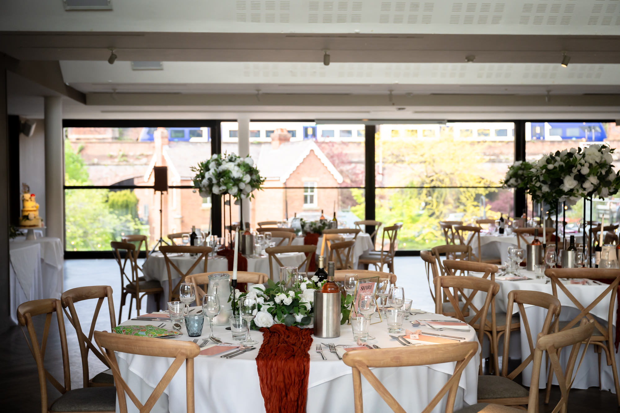 The Merchant Room at The Castlefield Rooms set up for the wedding breakfast, with round tables dressed in white linen, rust-coloured table runners, white floral centrepieces and views over Castlefield through the large windows.
