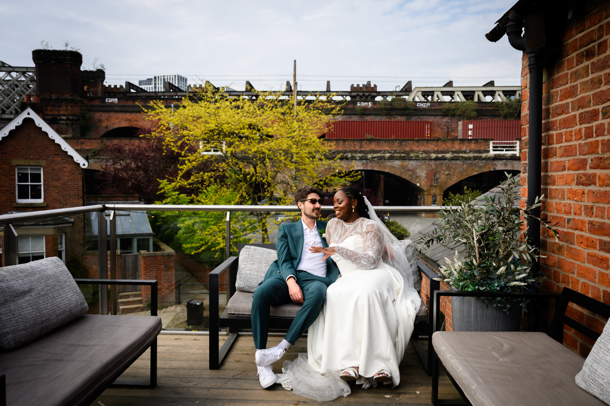 The bride and groom relaxing together on the balcony at The Castlefield Rooms while guests take their seats for the wedding breakfast, with the Castlefield viaduct and spring trees visible in the background.