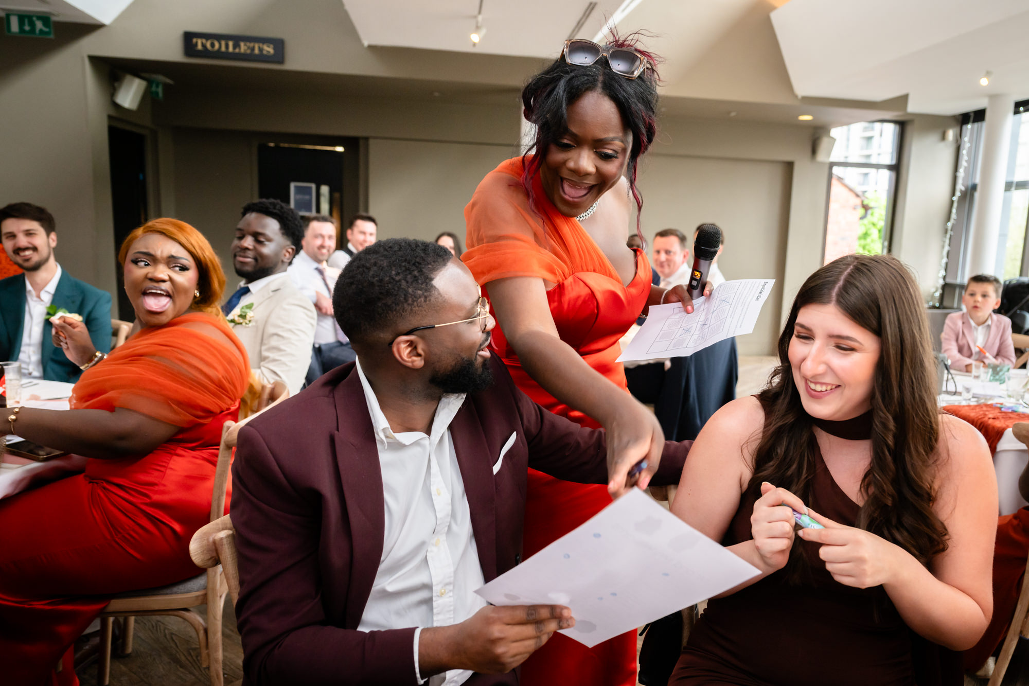 A wedding guest in an orange dress hosting a wedding game with a microphone during the wedding breakfast at The Castlefield Rooms, leaning over to show a guest their answer sheet.