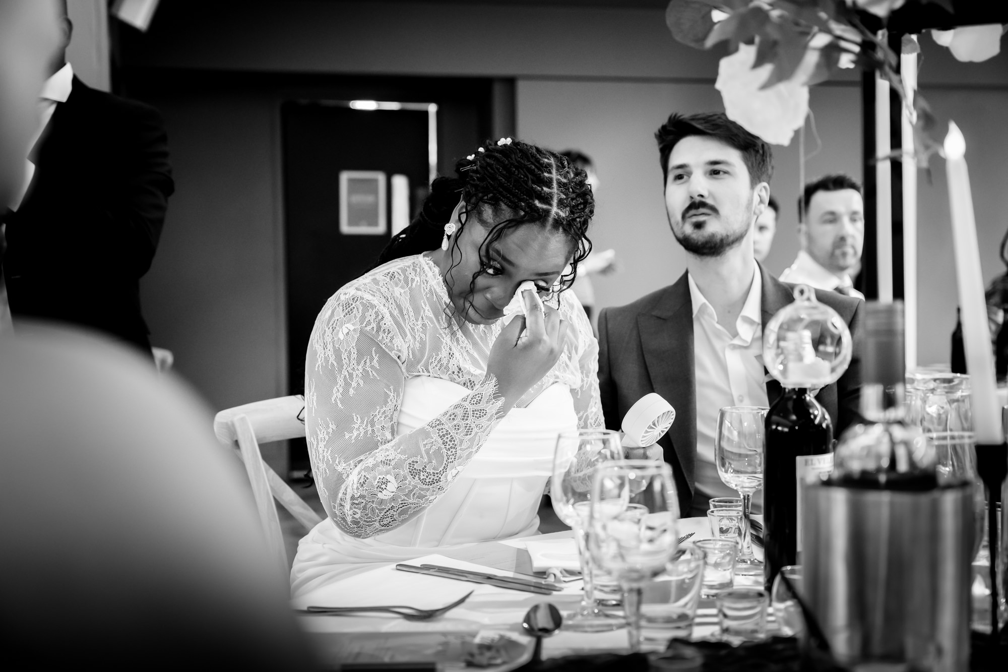 Black and white photo of the bride wiping a tear at the top table during speeches in the Merchant Room at The Castlefield Rooms, the groom watching beside her.