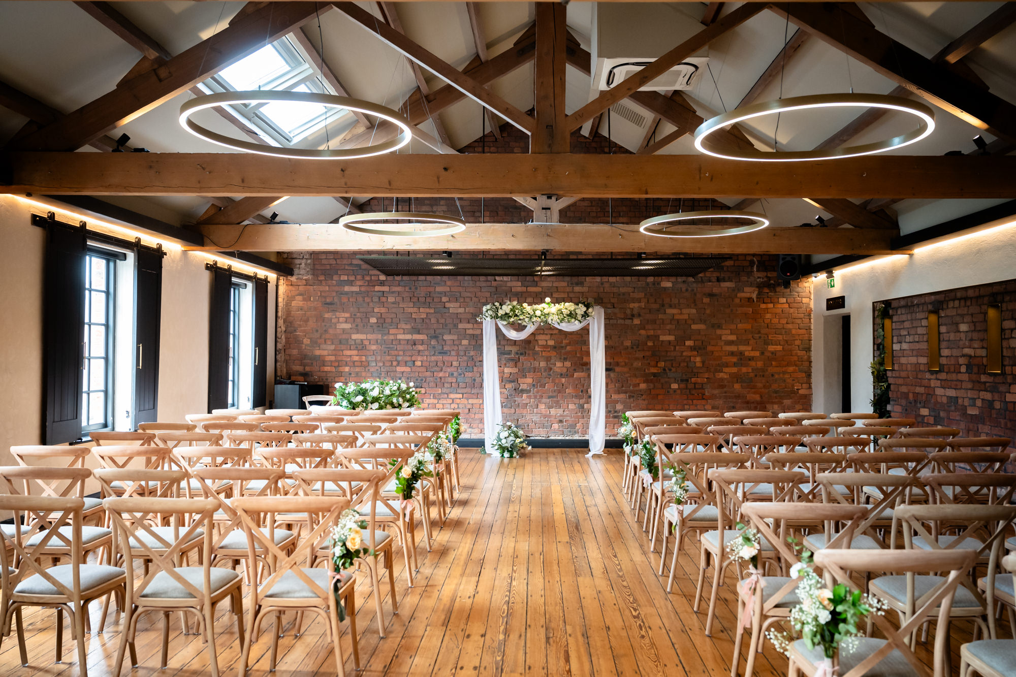 The Brindley Room at The Castlefield Rooms set up for a wedding ceremony, with wooden cross-back chairs lining the aisle, floral decorations and a white draped arch against the exposed brick wall.