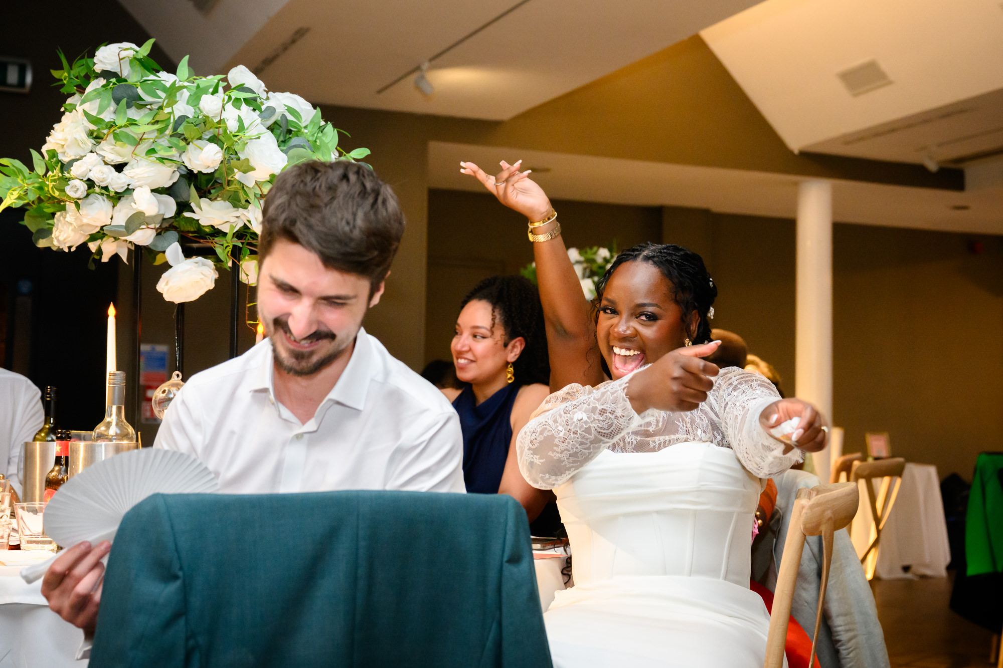 The bride pointing at a speaker during speeches at the top table in the Merchant Room at The Castlefield Rooms, the groom laughing beside her.