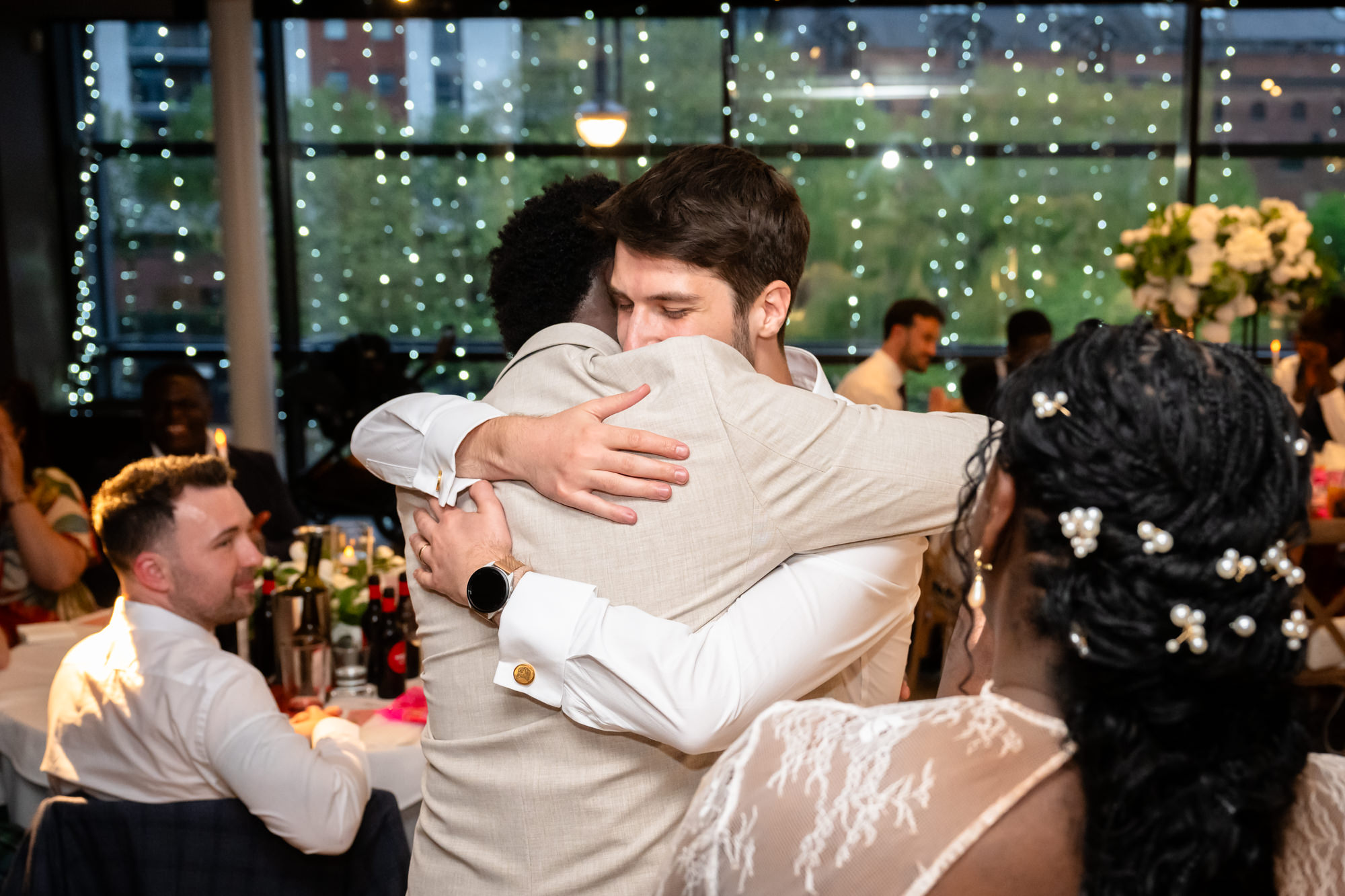 The groom hugging a male guest after speeches in the Merchant Room at The Castlefield Rooms, with the bride visible in the foreground and fairy lights in the windows behind.