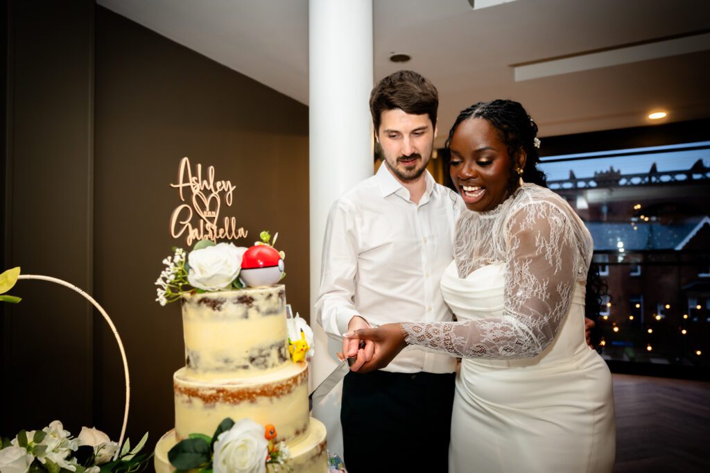 The bride and groom cutting their three-tiered wedding cake decorated with Pokemon figures and a personalised wooden topper reading Ashley and Gabriella at The Castlefield Rooms.