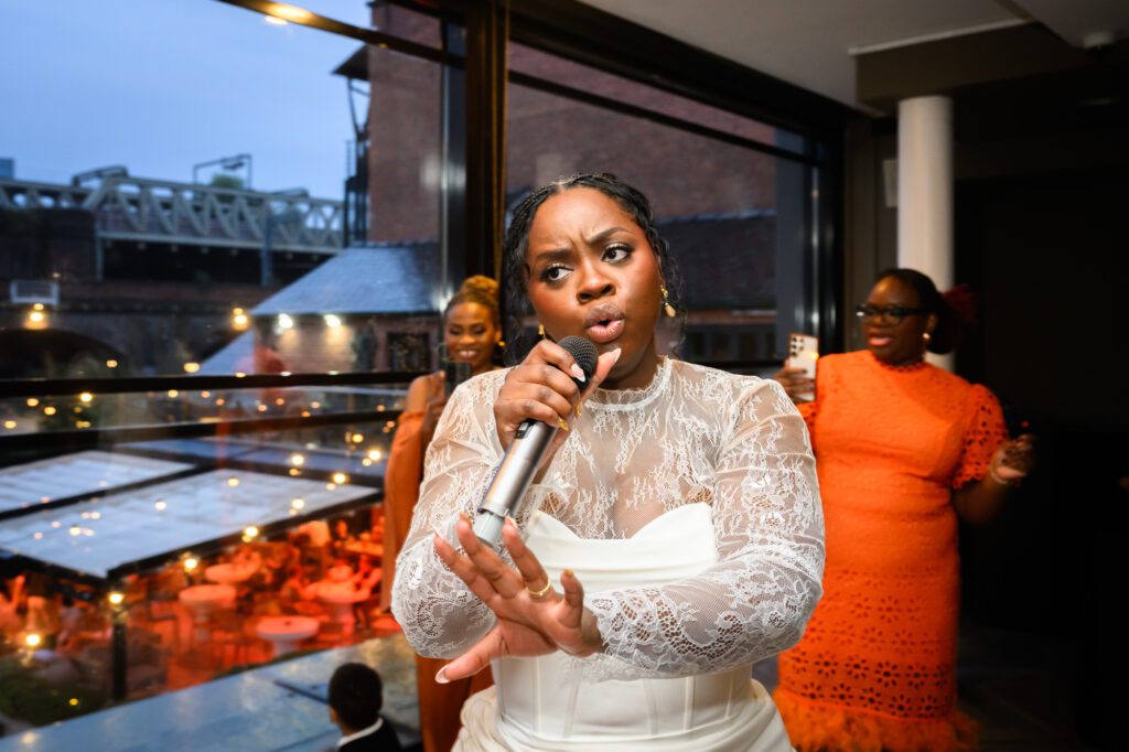 The bride singing into a microphone during the evening reception at The Castlefield Rooms, with the Castlefield viaduct lit up in the background through the windows.