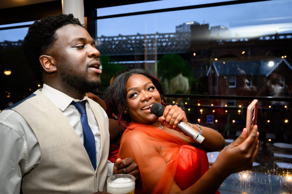 A wedding guest in an orange dress singing into a microphone with another guest during the evening reception at The Castlefield Rooms, with the Manchester skyline visible through the windows.