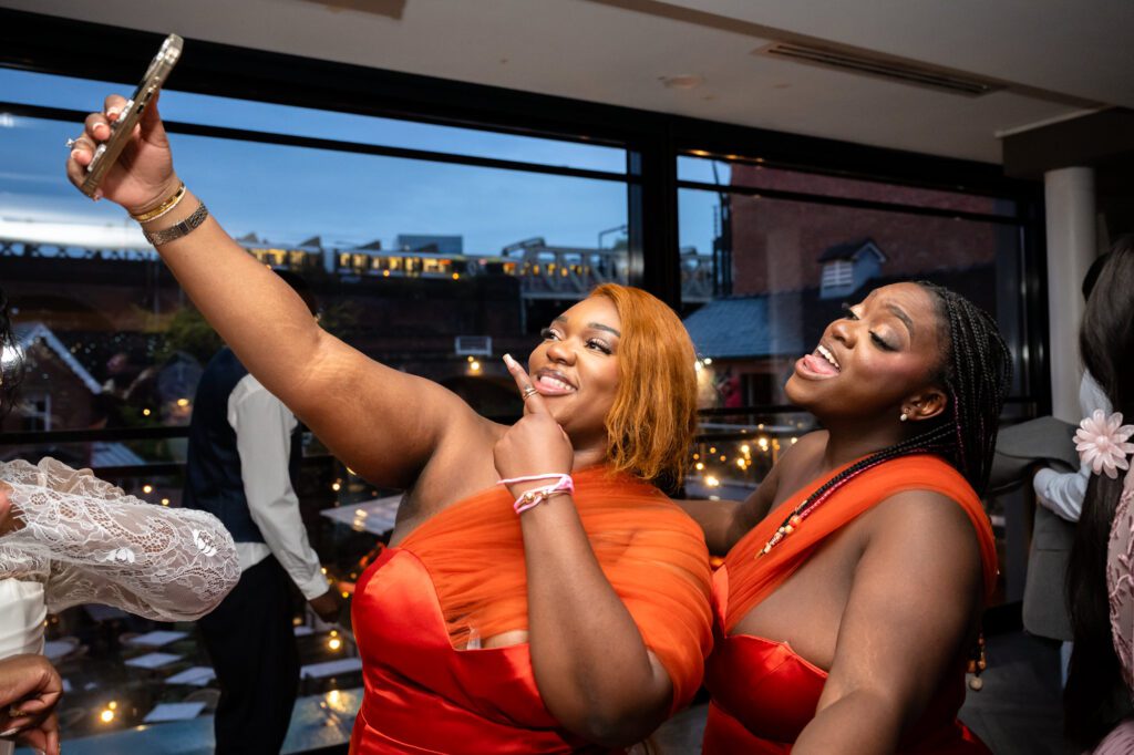 Two bridesmaids in orange dresses taking a selfie during the evening reception at The Castlefield Rooms, with the Castlefield skyline and fairy lights visible through the windows.