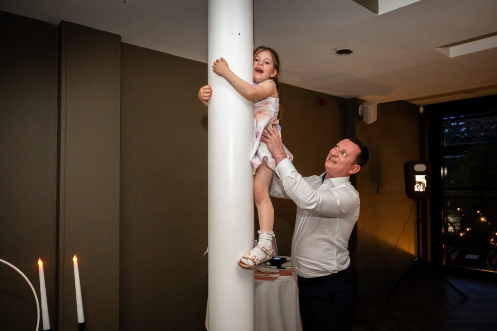 A young girl climbing a pillar in the Merchant Room at The Castlefield Rooms during the evening reception, helped by a wedding guest.
