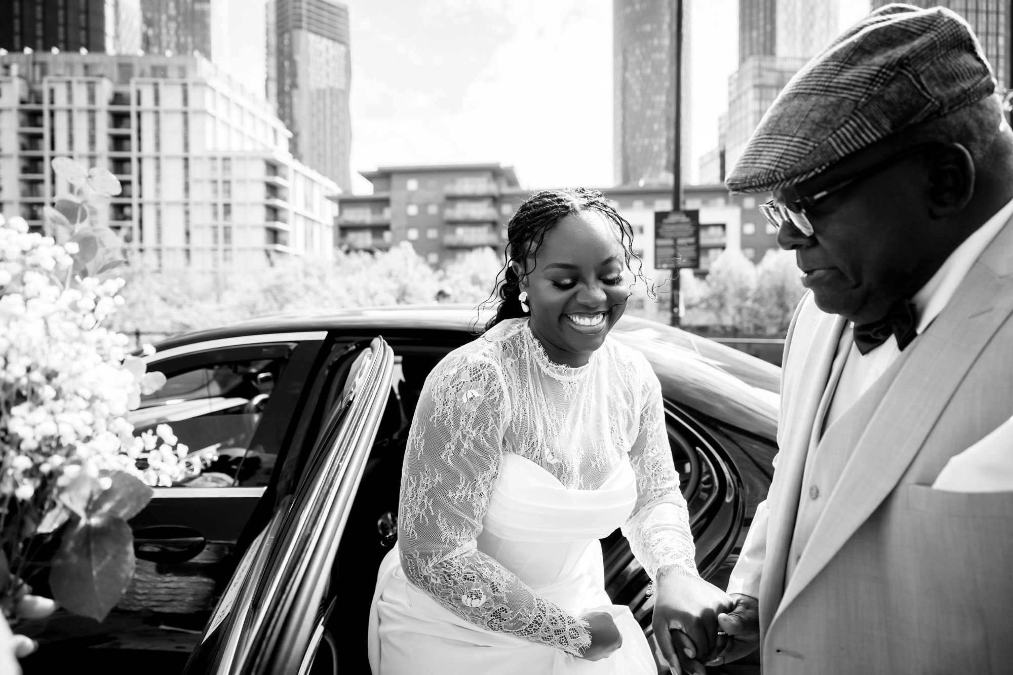 Black and white photo of the bride stepping out of a wedding car, smiling, with the Manchester city skyline in the background outside The Castlefield Rooms.