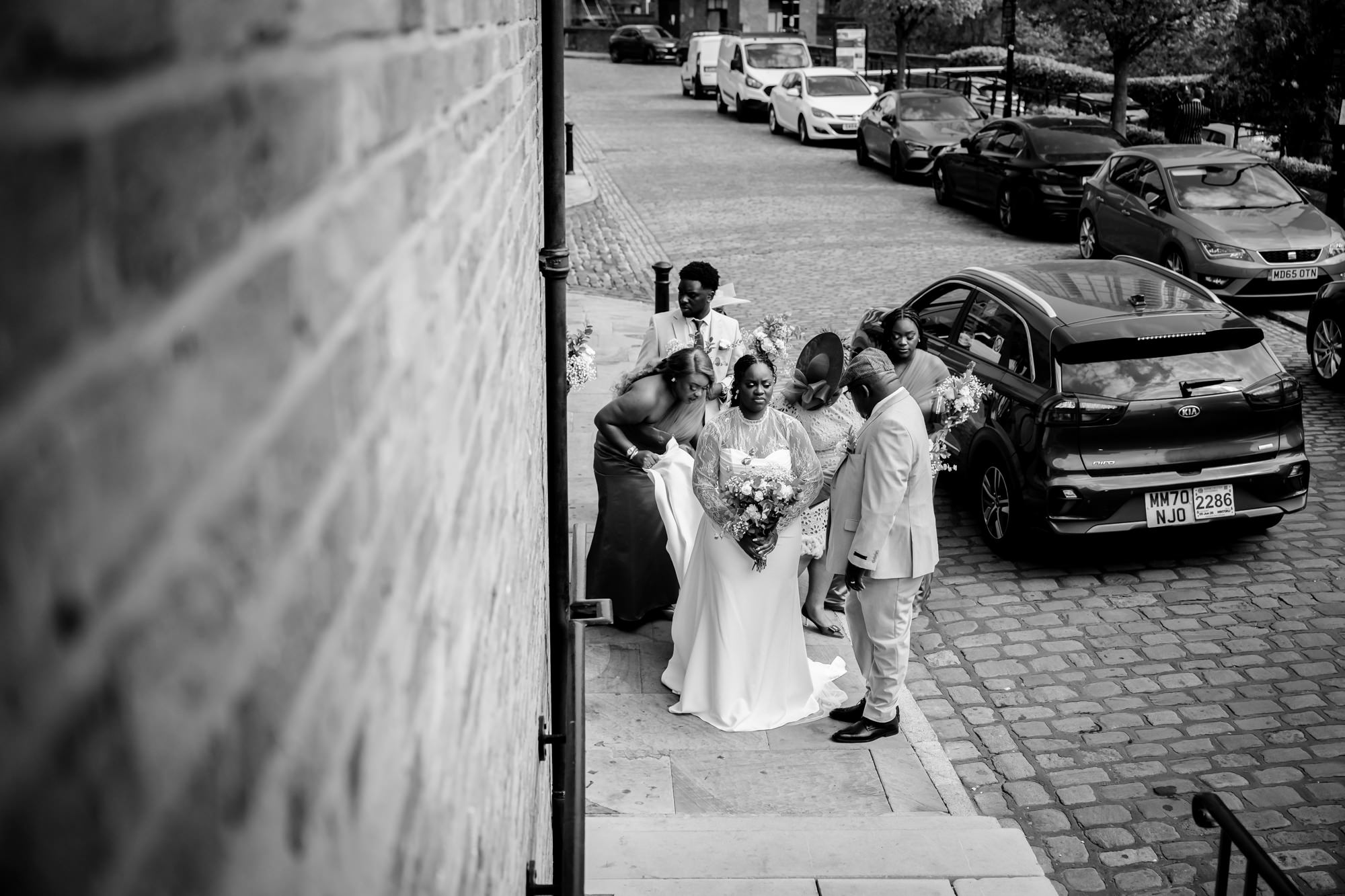Black and white aerial shot of the bride arriving outside The Castlefield Rooms, surrounded by bridesmaids and family on the cobbled street below.