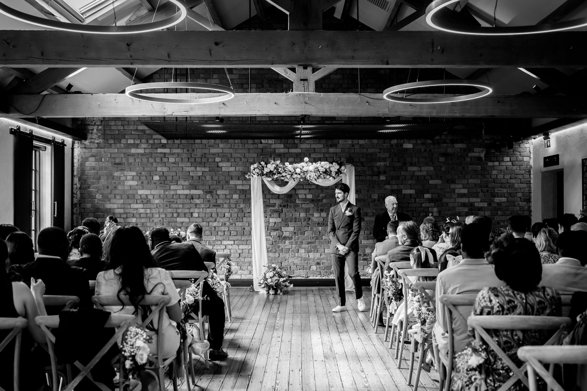 Black and white wide shot of the groom standing at the altar in the Brindley Room at The Castlefield Rooms, waiting for the bride with guests seated on either side.