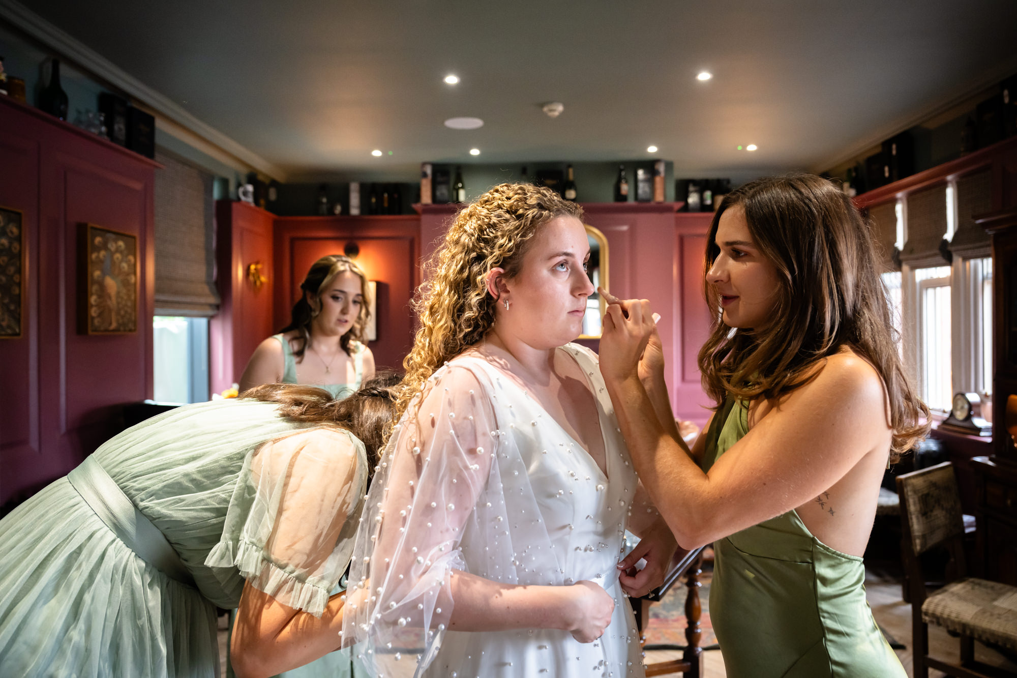 Bridesmaid applying makeup to the bride, with another bridesmaid in the background