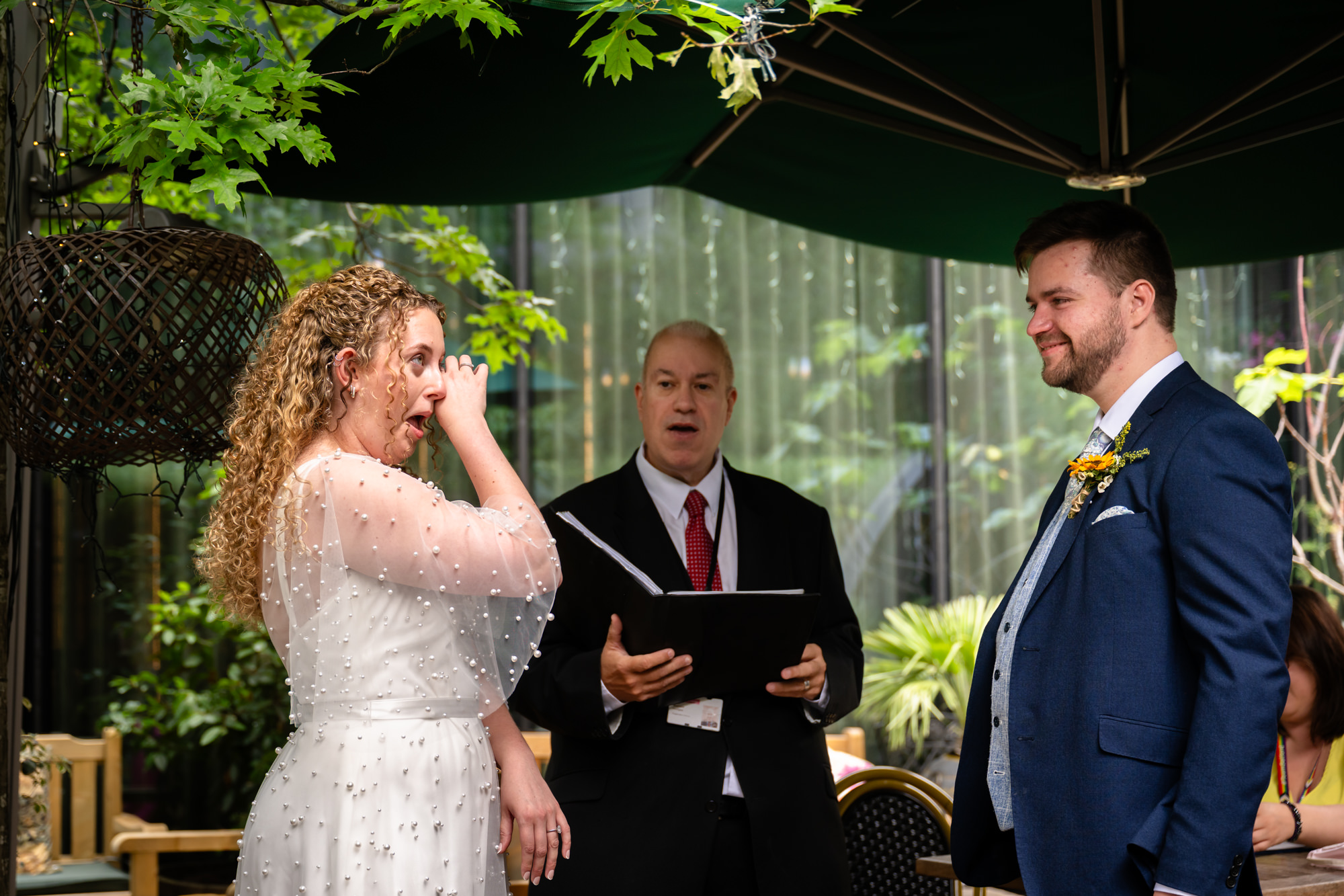 Bride wiping away a tear while laughing during the ceremony, celebrant reading in the background