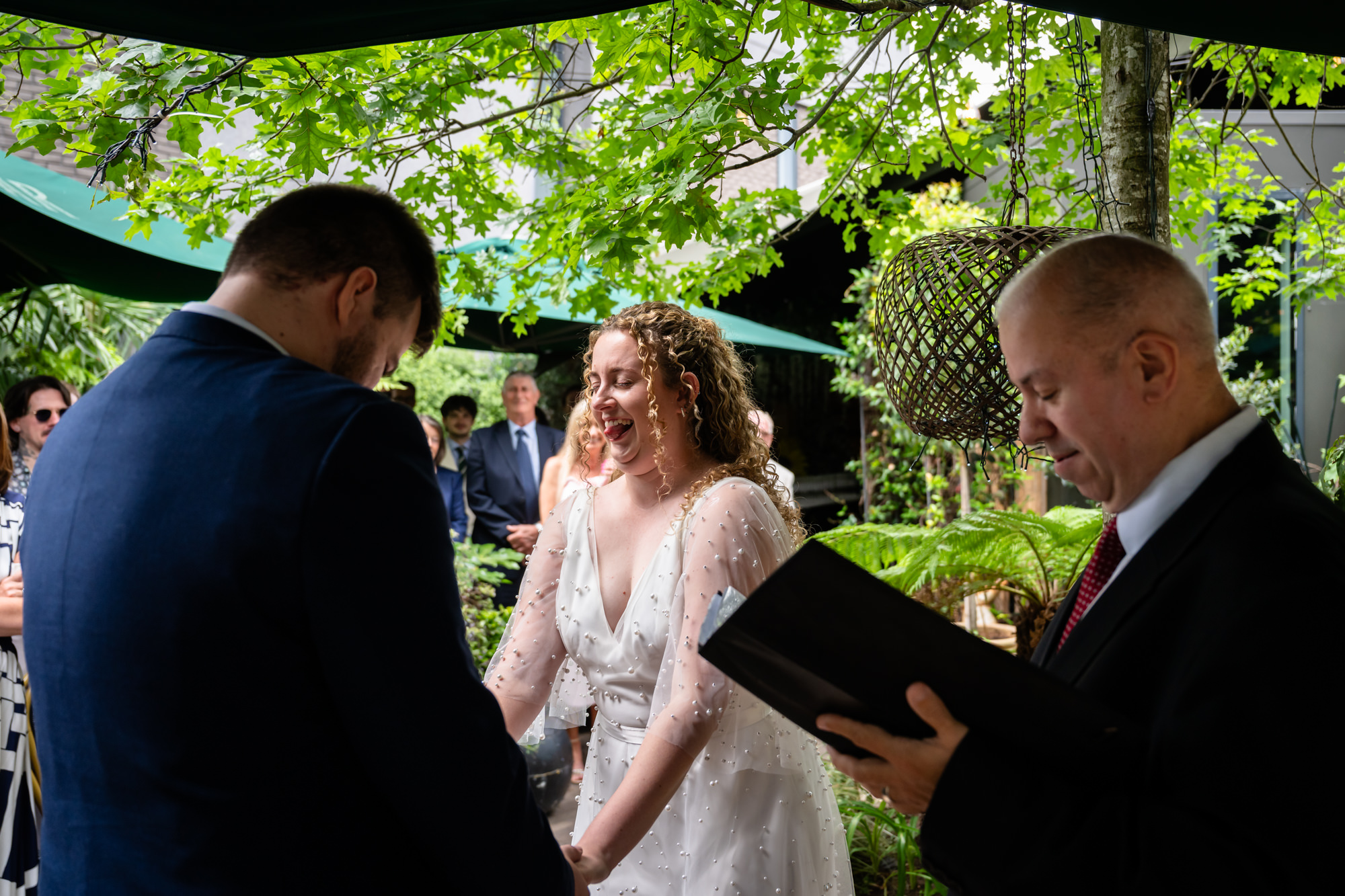 Bride laughing with her tongue out during the ceremony, groom looking down at his notes
