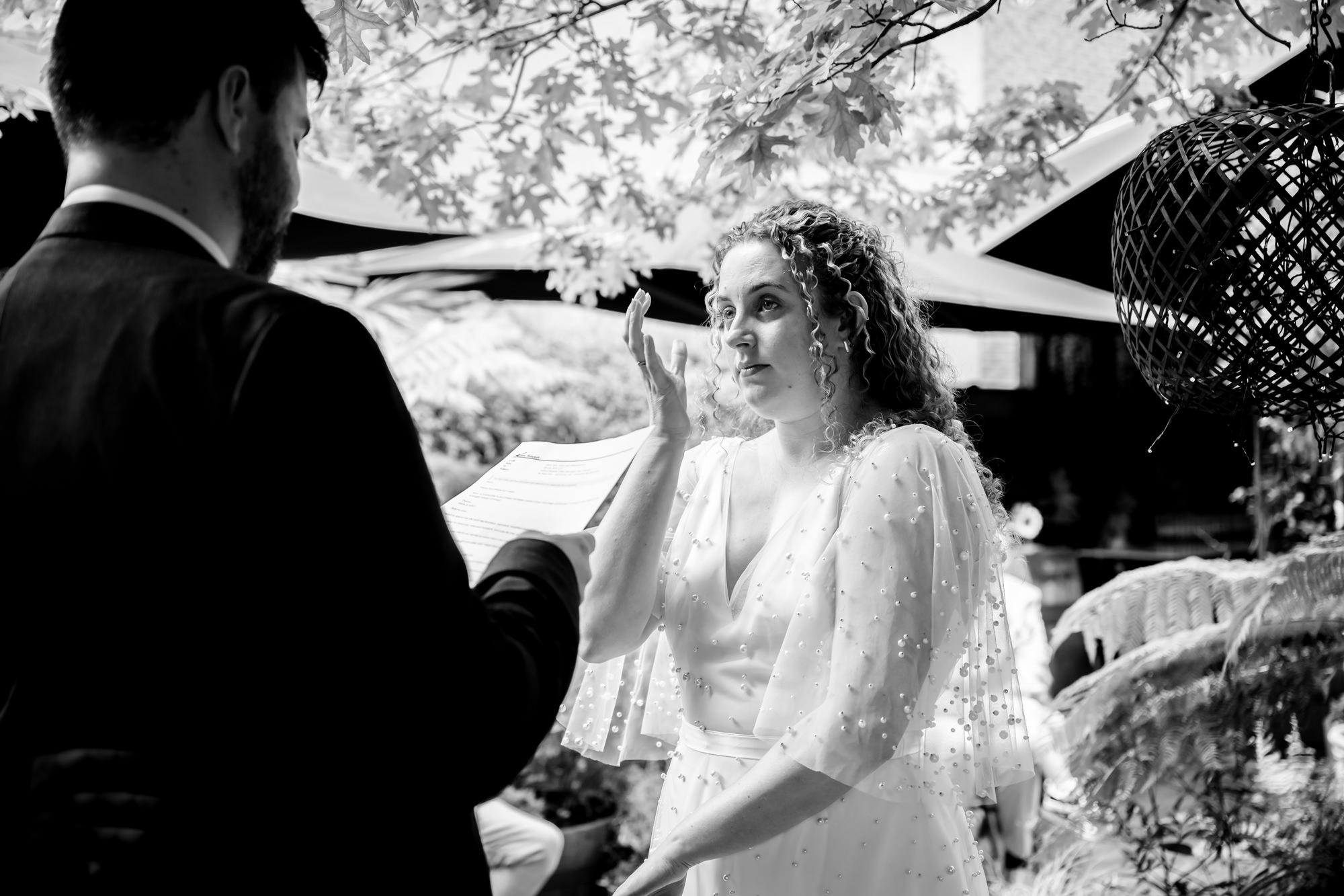 Bride reading her vows with a raised hand, groom listening, black and white