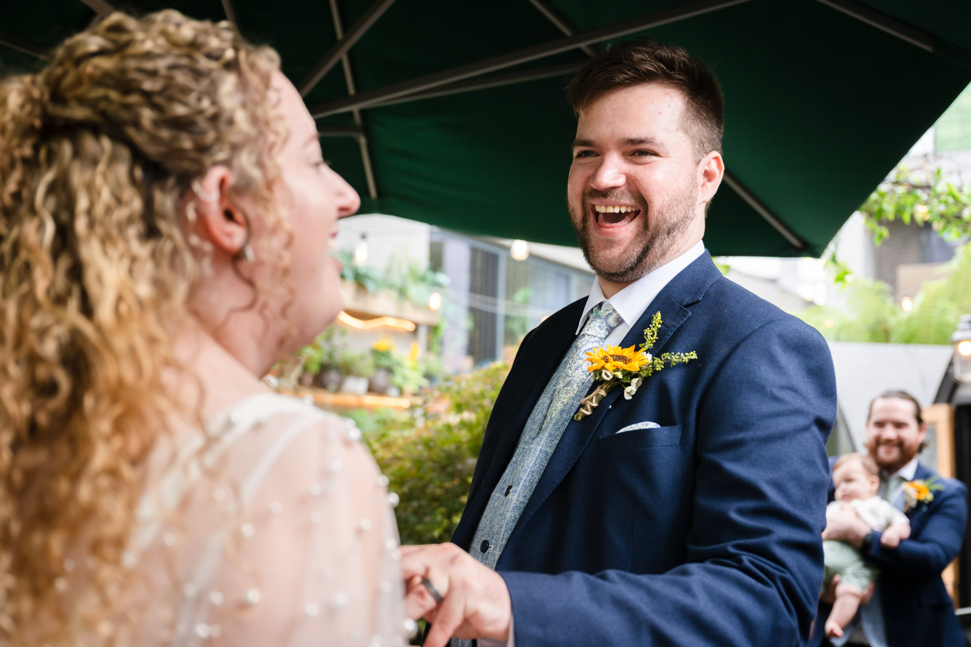 Groom laughing while holding the bride's hand during the ceremony, a baby visible among the guests
