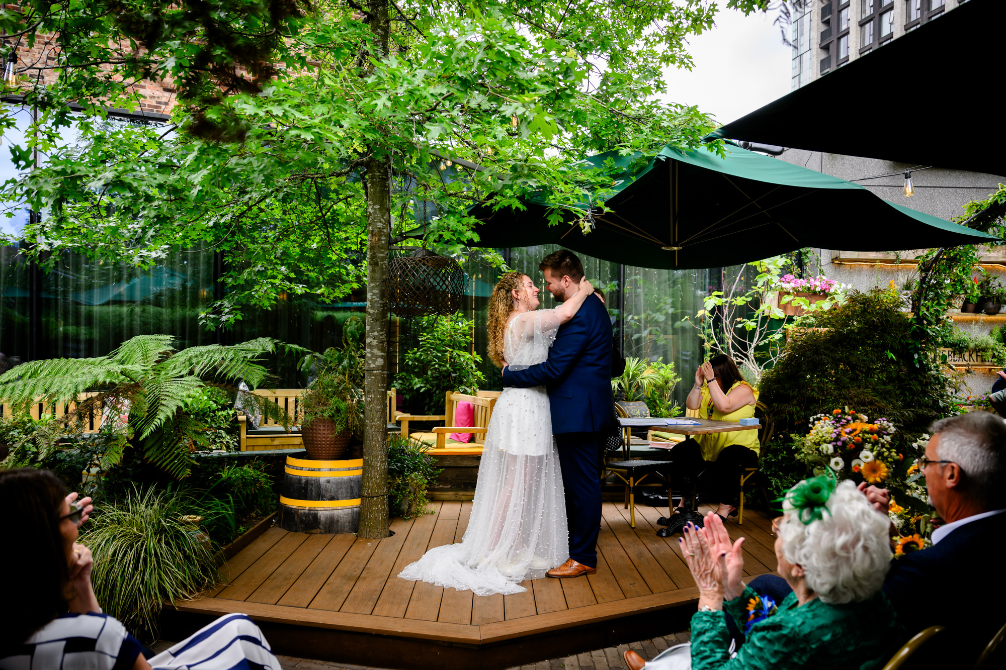 Newlyweds embracing after the ceremony on a wooden deck, guests clapping in the garden