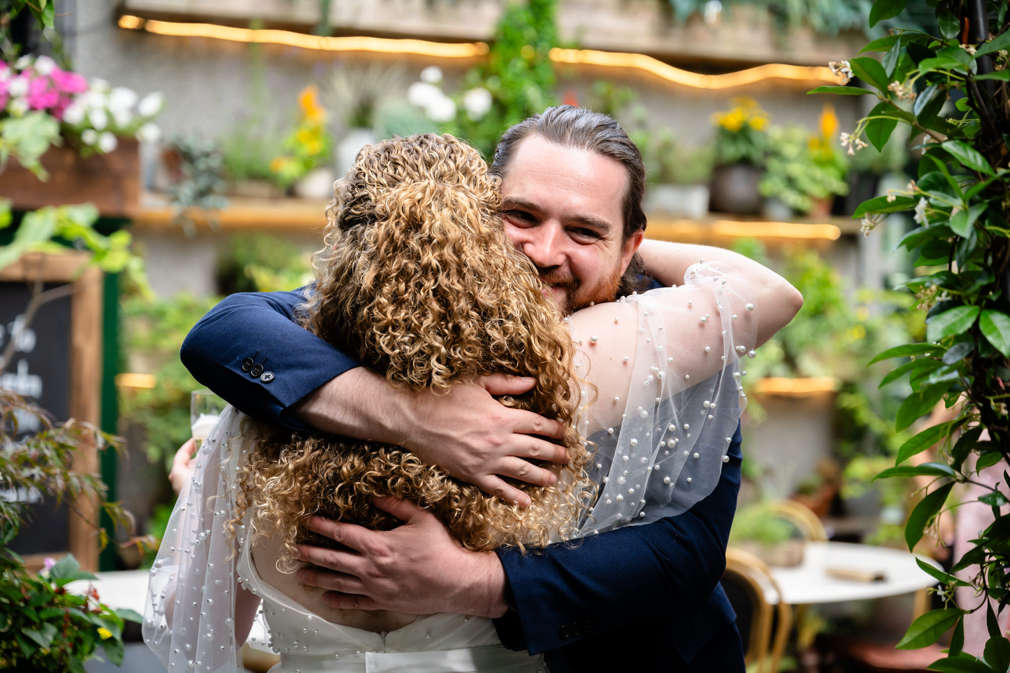 Bride and groom embracing at the drinks reception, guests and greenery in the background