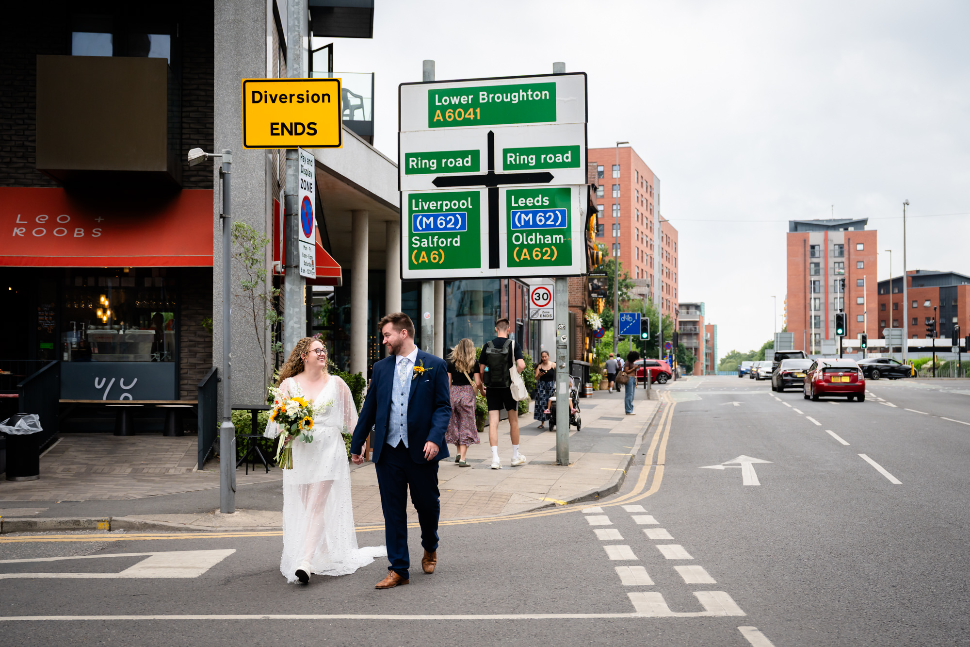Bride and groom walking hand in hand past road signs for Salford near the Black Friar