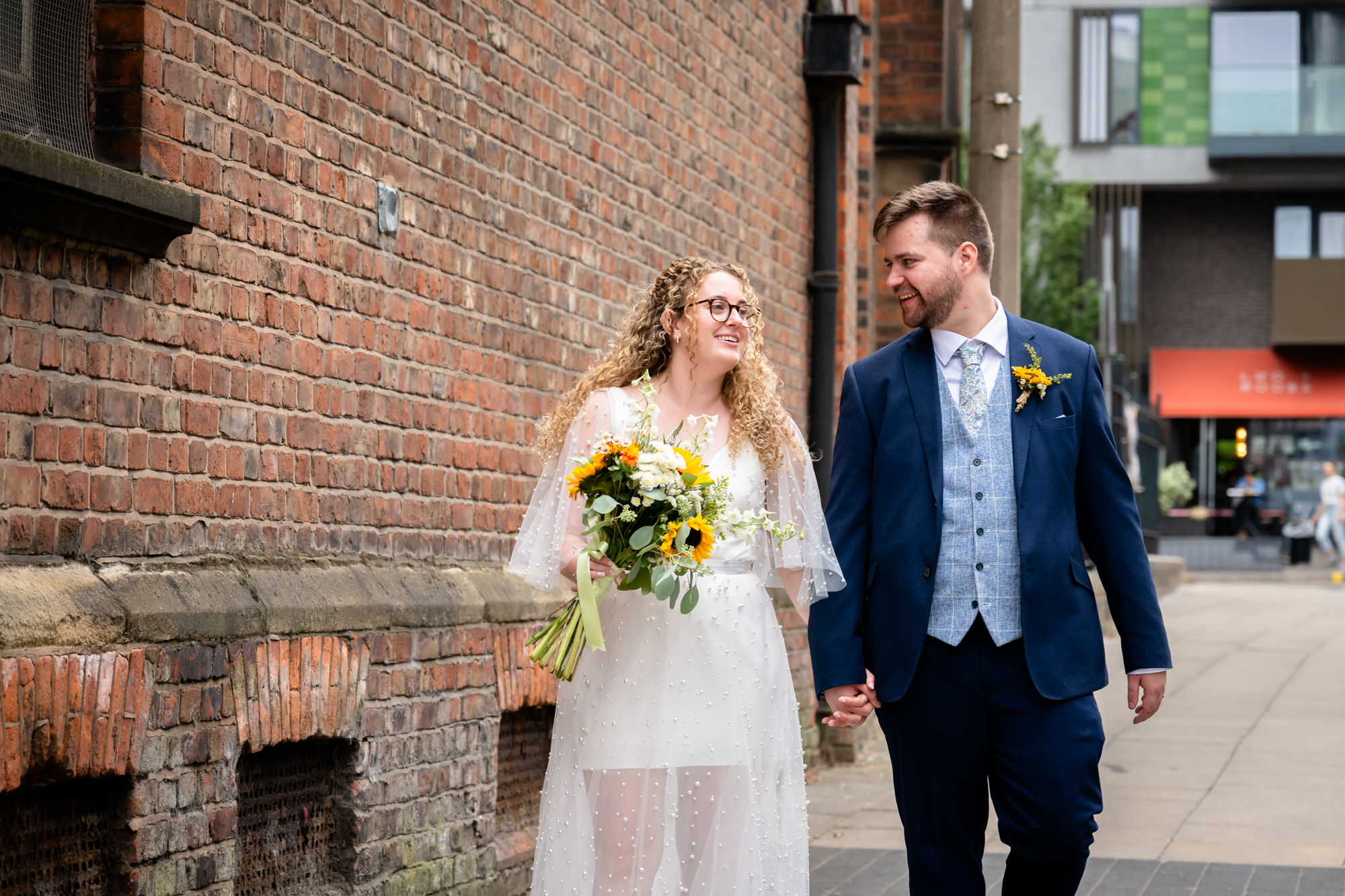 Bride and groom walking past a brick wall, bride holding a sunflower bouquet