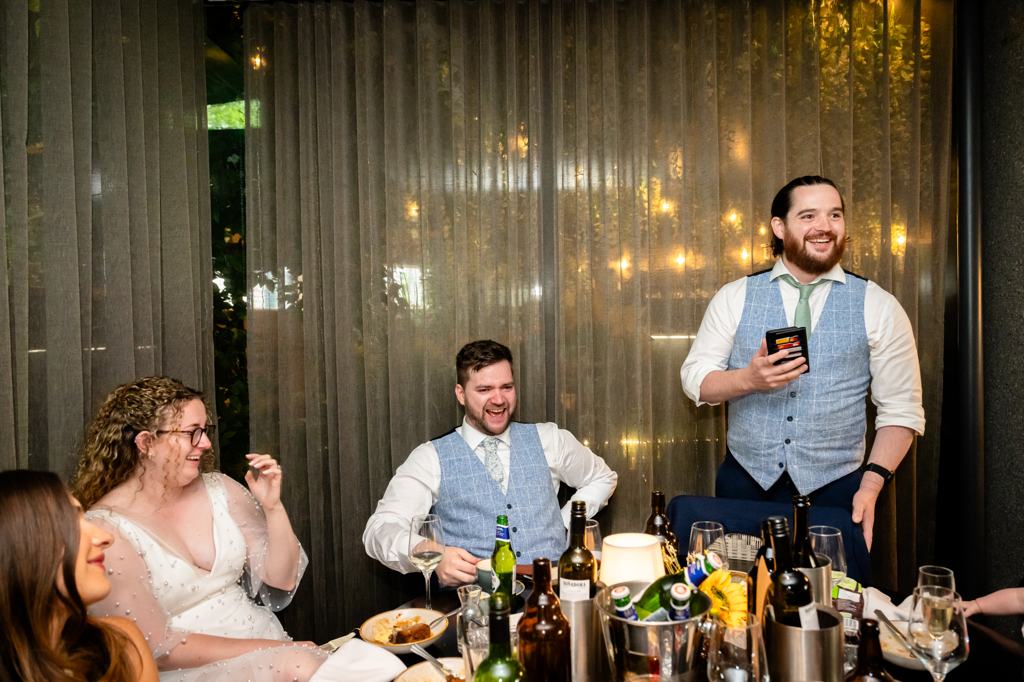 Wedding guest giving a speech, holding notes, bride and groom laughing beside him