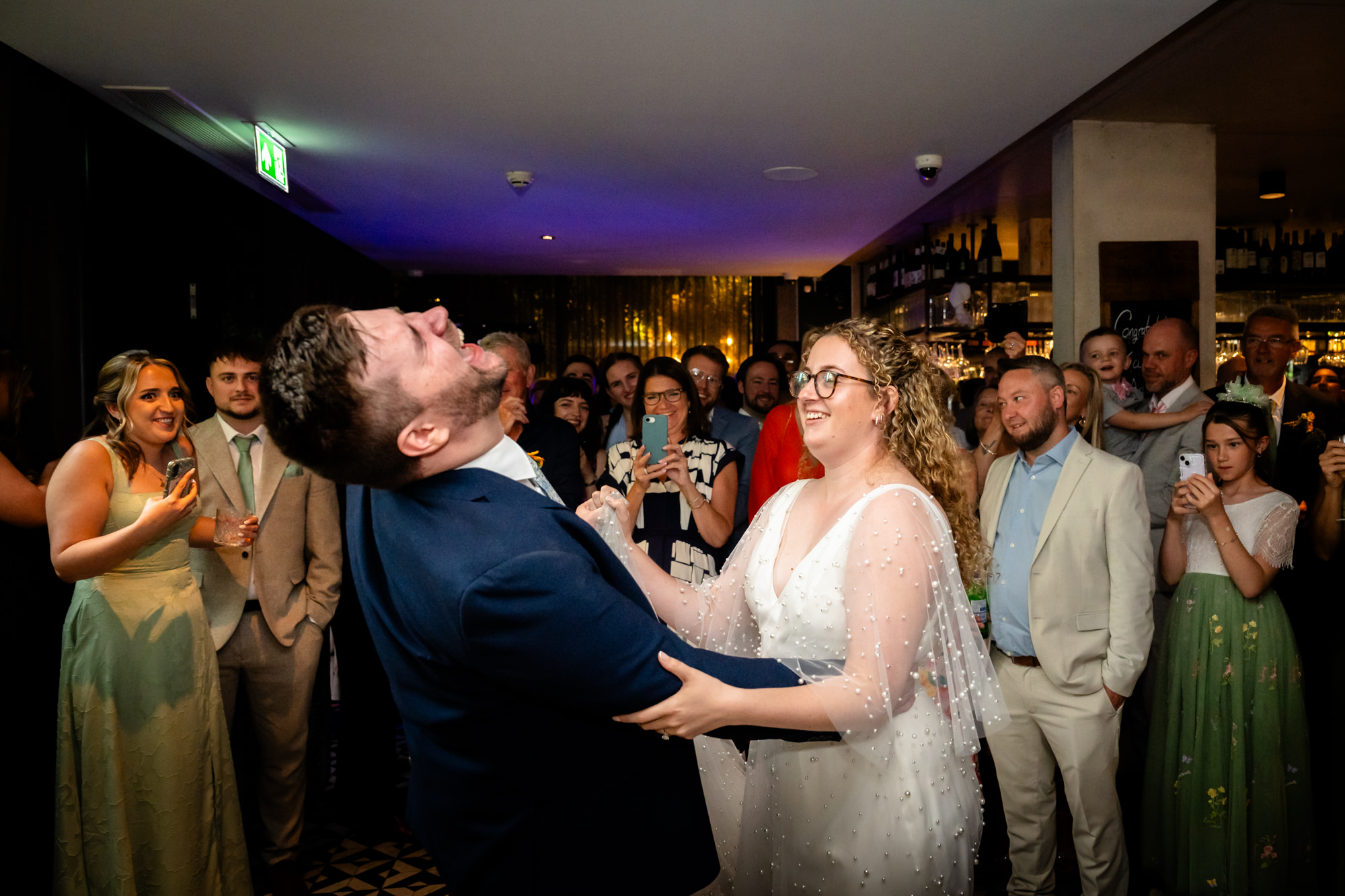 Groom throwing his head back laughing while dancing with the bride, guests watching