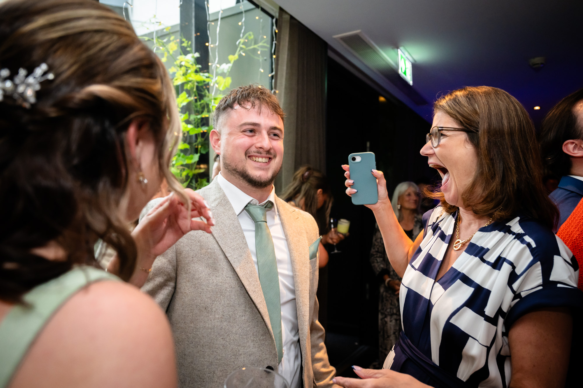 Wedding guest laughing while another takes a photo on her phone