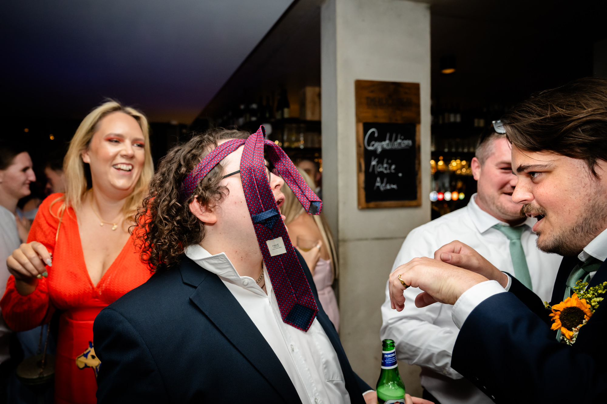 Wedding guest with a tie tied around his head, friends laughing beside a Congratulations Katie and Adam chalkboard